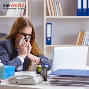 Getting your flu and COVID-19 vaccinations together: why it's safe and important. Image depicts a woman at her desk in the office blowing her nose.